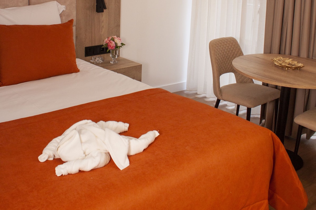 A hotel room with a large bed, a bedside table with a vase of flowers, a round table with chairs, and a window with sheer curtains.