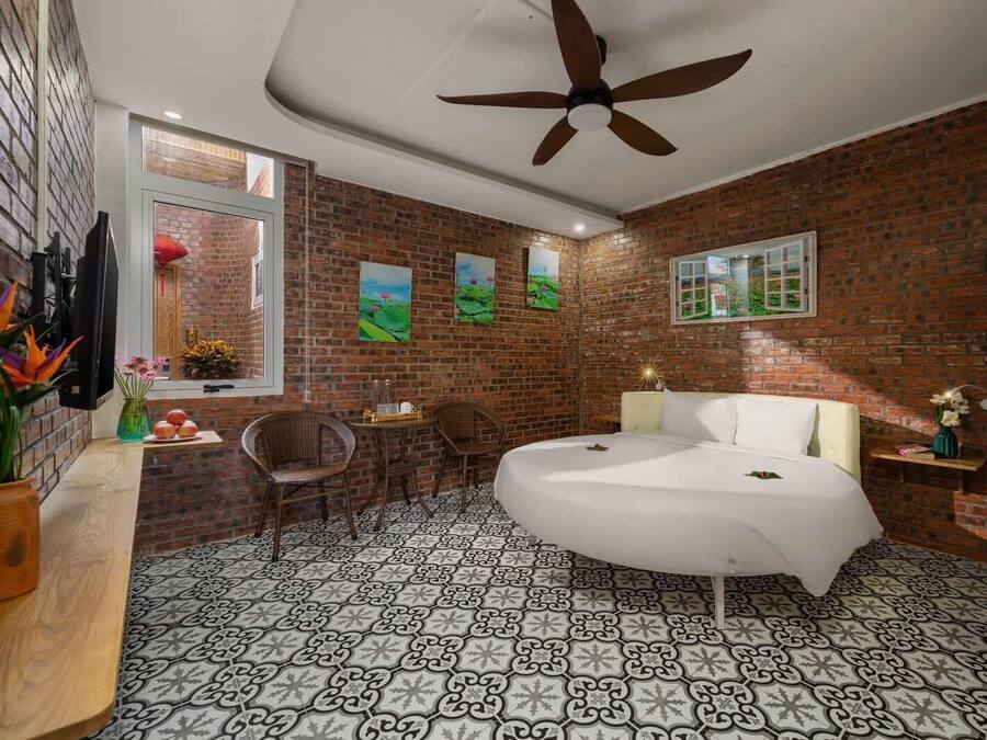 A bedroom with a brick wall, a round bed, a ceiling fan, and a window with artwork.