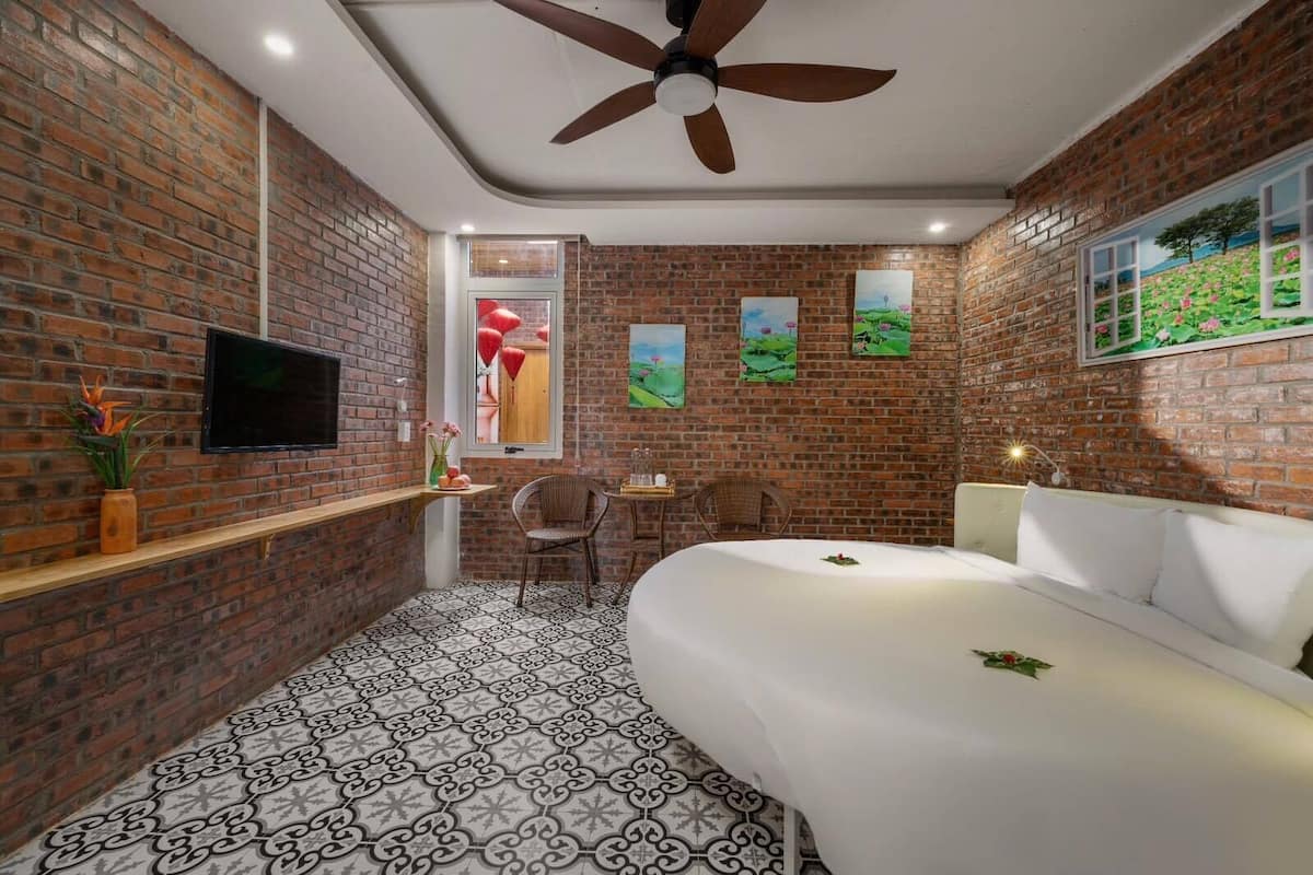 A bedroom with a brick wall, a ceiling fan, a bed, a TV, and a window with a painting.