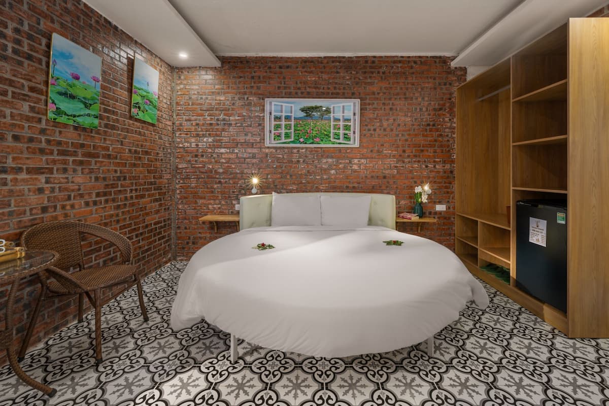 A room with a brick wall, a bed with white bedding, a wicker chair, a small table, and a wooden cabinet.
