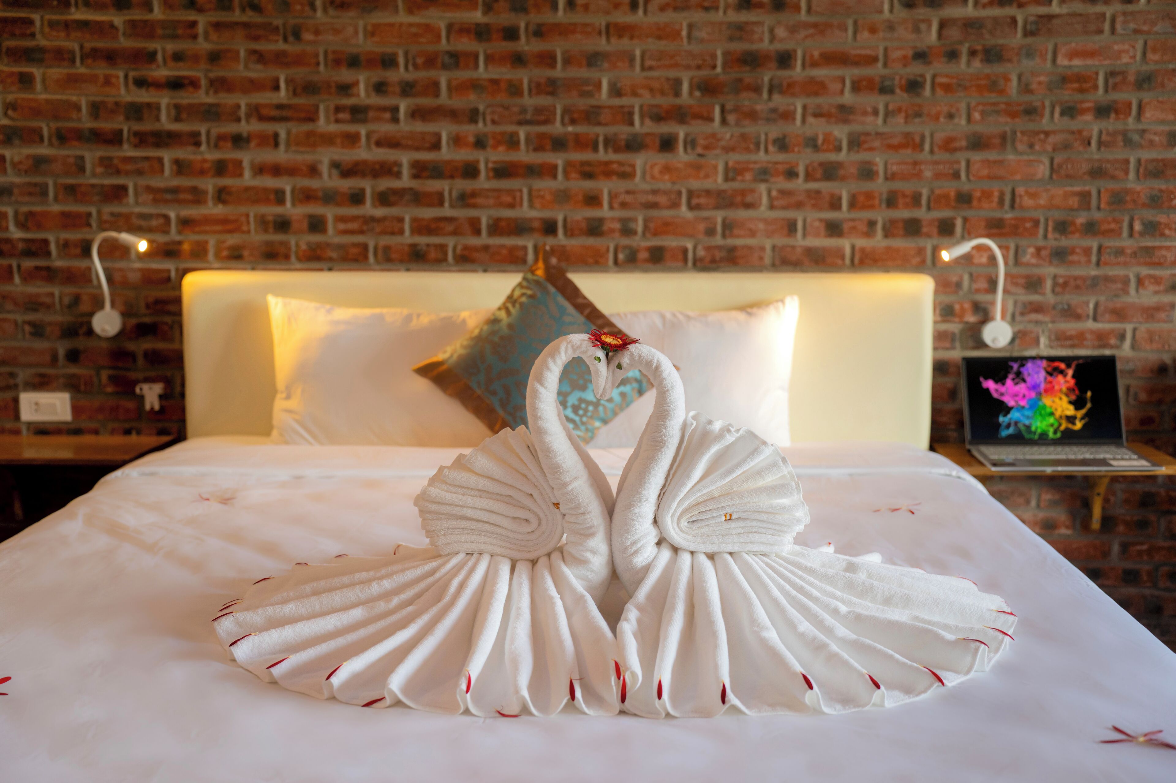 A hotel room with a brick wall, a neatly made bed with white bedding, and a laptop on a side table.