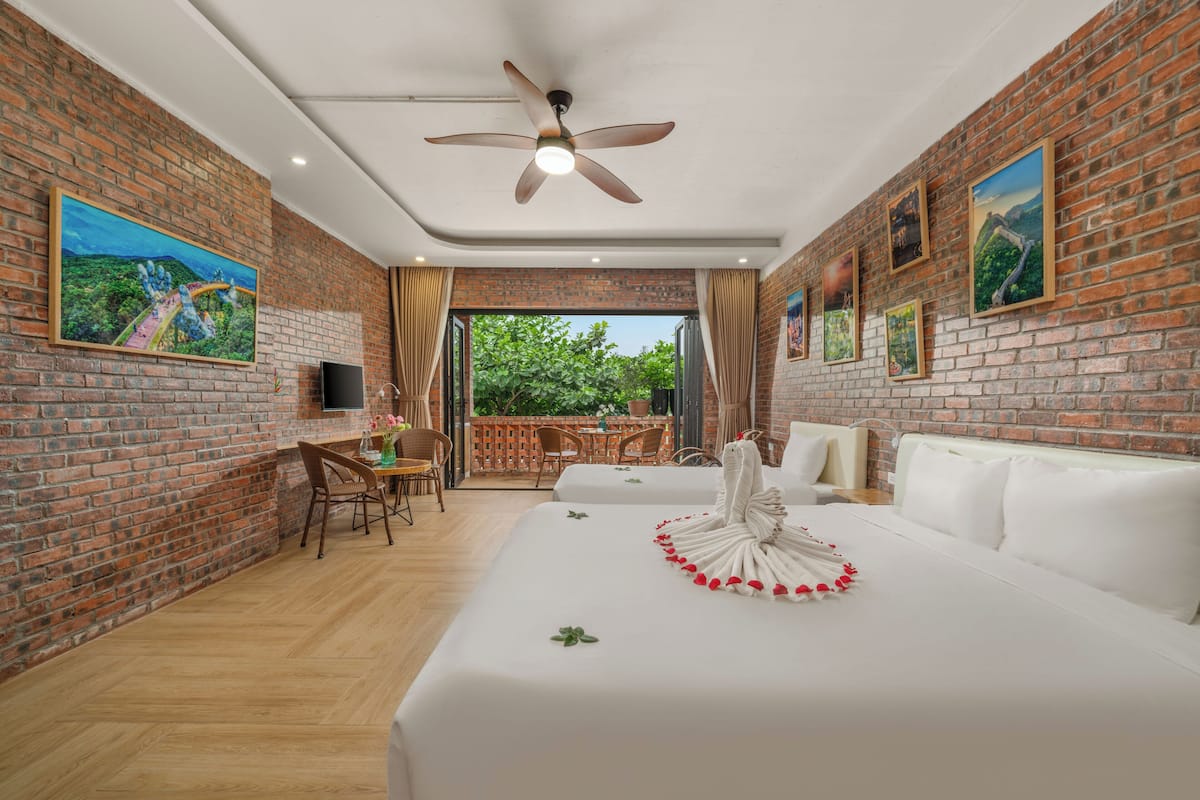 A hotel room with a brick wall, a ceiling fan, a bed with white bedding, a dining area with a table and chairs, and a view through a glass door.