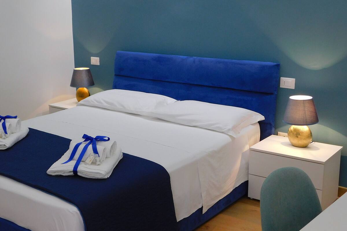 Een keurig opgemaakt bed met blauw beddengoed, twee handdoeken met blauwe linten, een bedlamp en een klein tafeltje met een lamp.