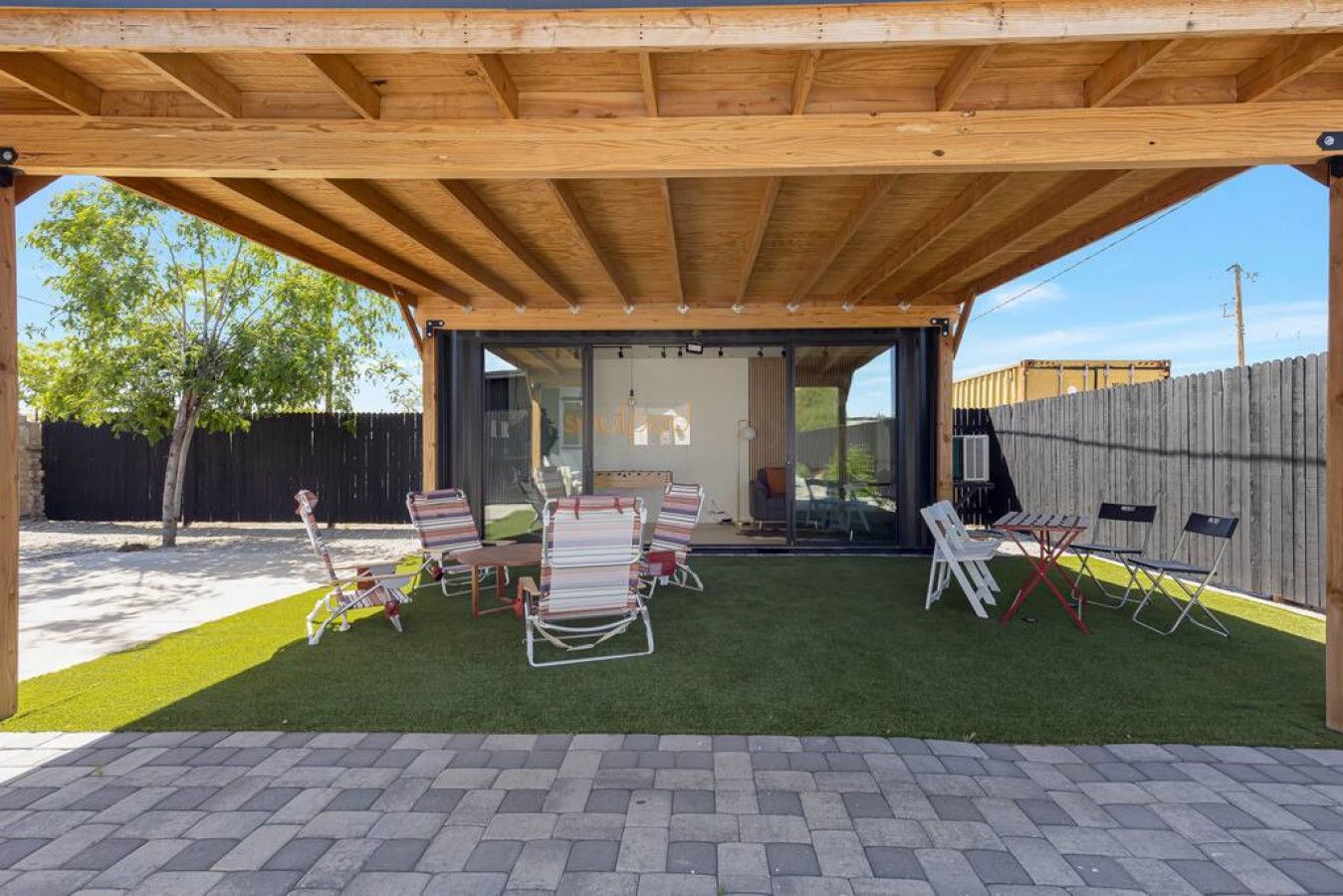 An outdoor patio with a wooden pergola, a seating area with chairs and tables, and a view of a house through a glass door.