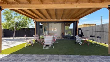 An outdoor patio with a wooden pergola, a seating area with chairs and tables, and a view of a house through a glass door.