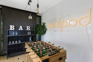 Una habitación con mesa de fútbol, zona de bar con estanterías y una pared con la palabra 'soulpod' en letras amarillas.