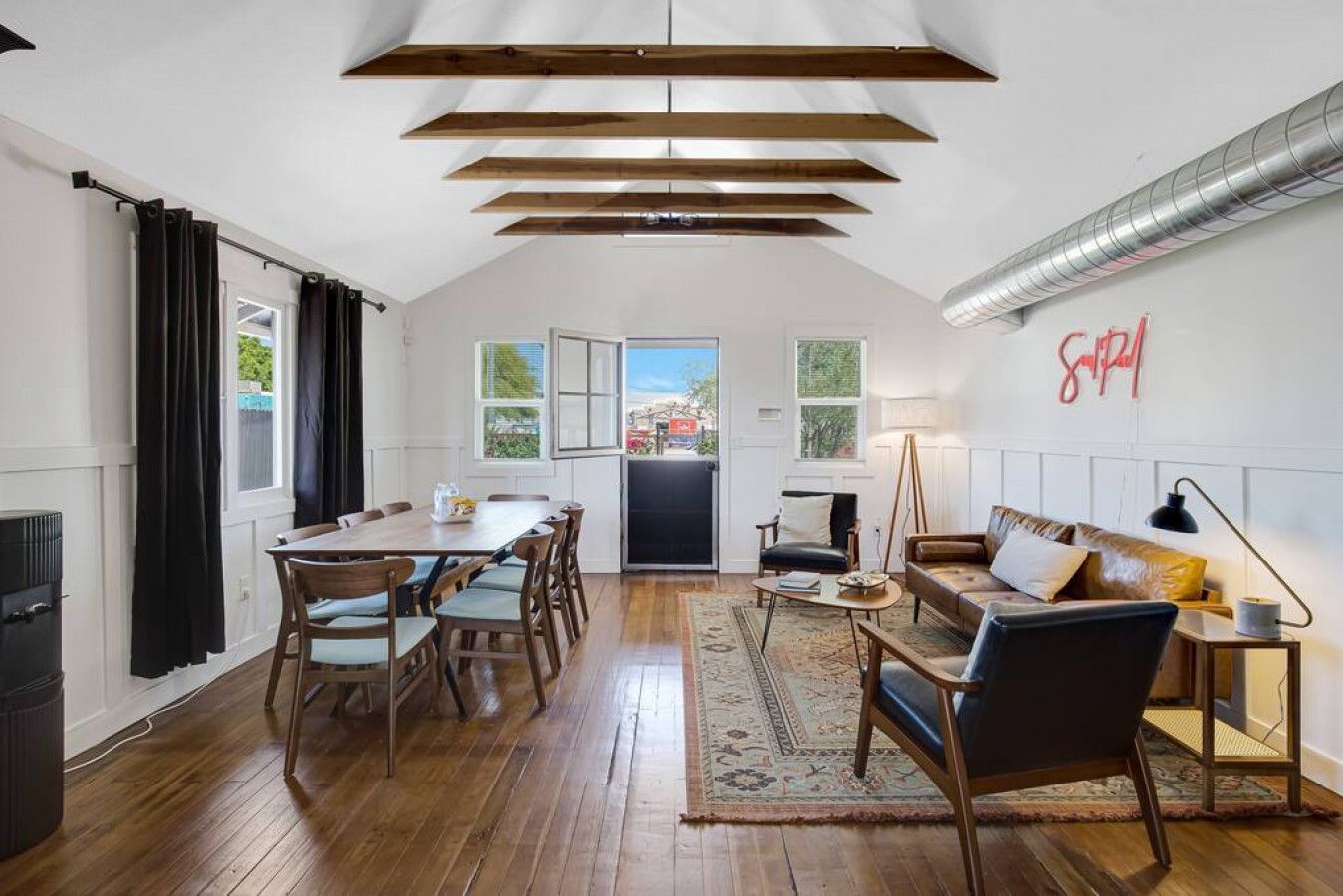 A spacious living area with a dining table, chairs, a sofa, and a coffee table. There is a large window with curtains, a door leading outside, and a wall-mounted radiator.