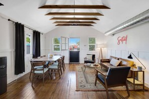 Amplia sala de estar con mesa de comedor, sillas, sofá y mesa de centro. Hay un ventanal con cortinas, una puerta que da al exterior y un radiador fijado a la pared.