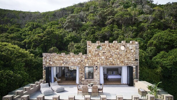 Une structure en pierre qui ressemble à un château, dotée d’un balcon, de sièges confortables et offrant une vue sur une végétation luxuriante.