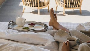 Un balcon avec deux chaises en bois, un plateau contenant une théière, des tasses et des fruits, et un lit où une personne se détend.