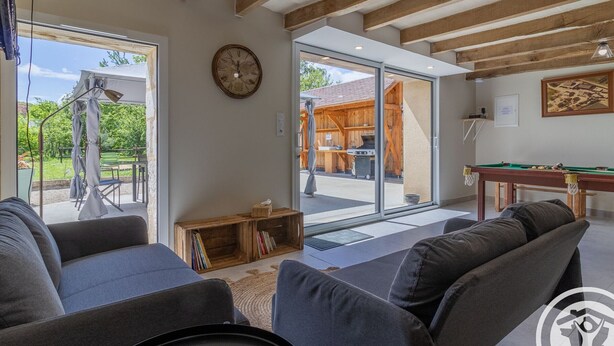 A modern living room with grey sofas, a wooden coffee table, a pool table, and a large window with a view of an outdoor patio. - Spacious Retreat: 5BR, Games Room, Forest Edge, and Large Garden Close to Château and Zoo (Baugé en Anjou)