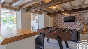 A cozy indoor space with a foosball table, a bar area, and a television mounted on a leaf-patterned wall. - Spacious Retreat: 5BR, Games Room, Forest Edge, and Large Garden Close to Château and Zoo (Baugé en Anjou)