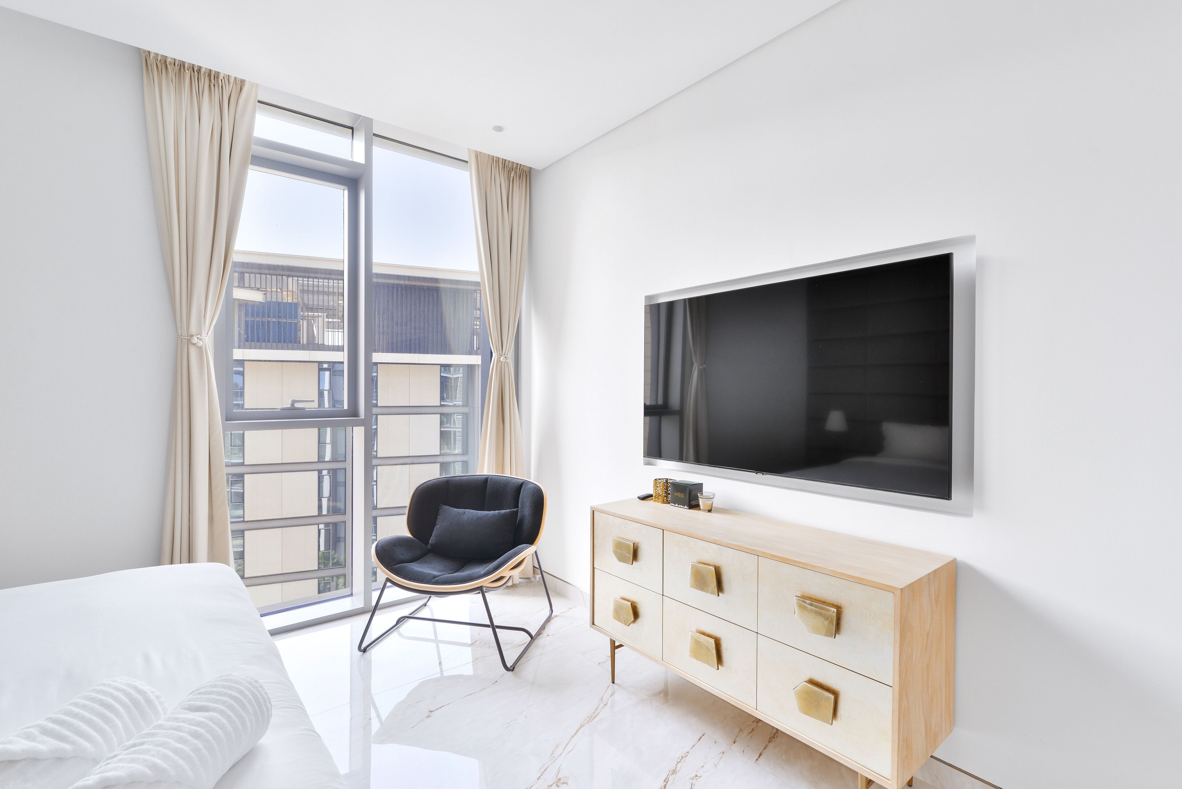 Quarto de hotel moderno com uma janela grande, televisão de tela plana, cômoda de madeira e uma cadeira preta.