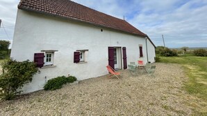 A white building with a red-tiled roof, two windows with shutters, and a gravel driveway.