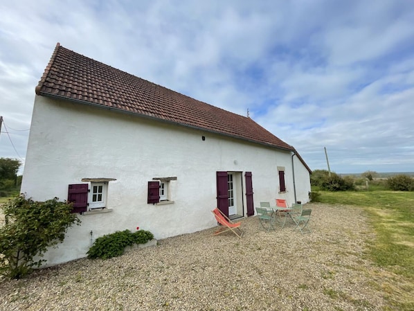 A white building with a red-tiled roof, two windows with shutters, and a gravel driveway.
