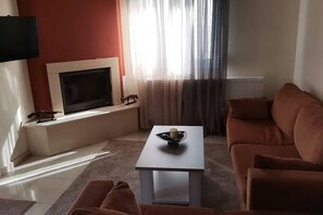 A living room with a flat-screen TV mounted on a wall, a sofa, a coffee table, and a window with sheer curtains. - Sweet Little Home Trikala (Pyli)