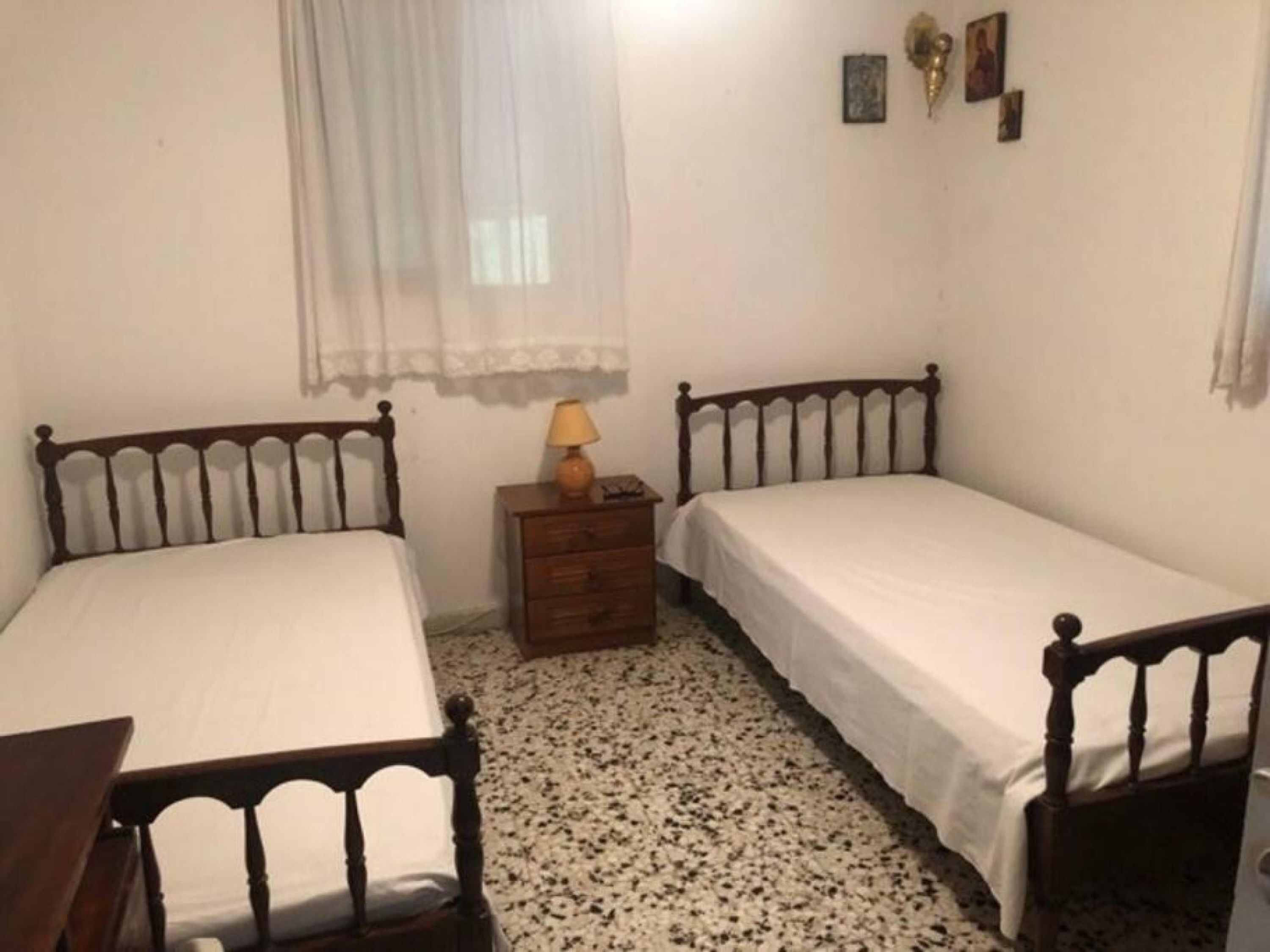 A small, simple room with two single beds, a wooden nightstand, a lamp, and a small dresser.