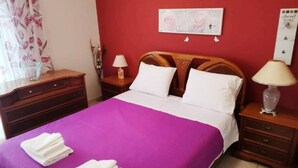 A hotel room with a bed, two nightstands, a wooden headboard, a wall with a floral painting, and a window with curtains. - Alex s Apartment in Trikala (Trikala)
