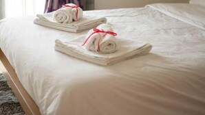 A neatly made bed with white linens and two rolled towels tied with red ribbons. - Dimsim Home- House With Name (Trikala)