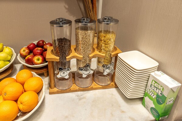 A kitchen counter with food dispensers, a bowl of apples, a bowl of oranges, and a carton of milk.