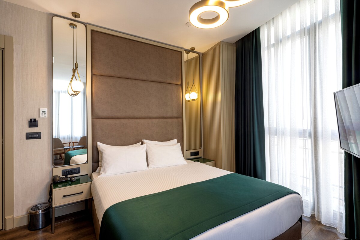 Camera d'albergo con un letto grande, uno specchio, un comodino, una televisione e una finestra con tende.