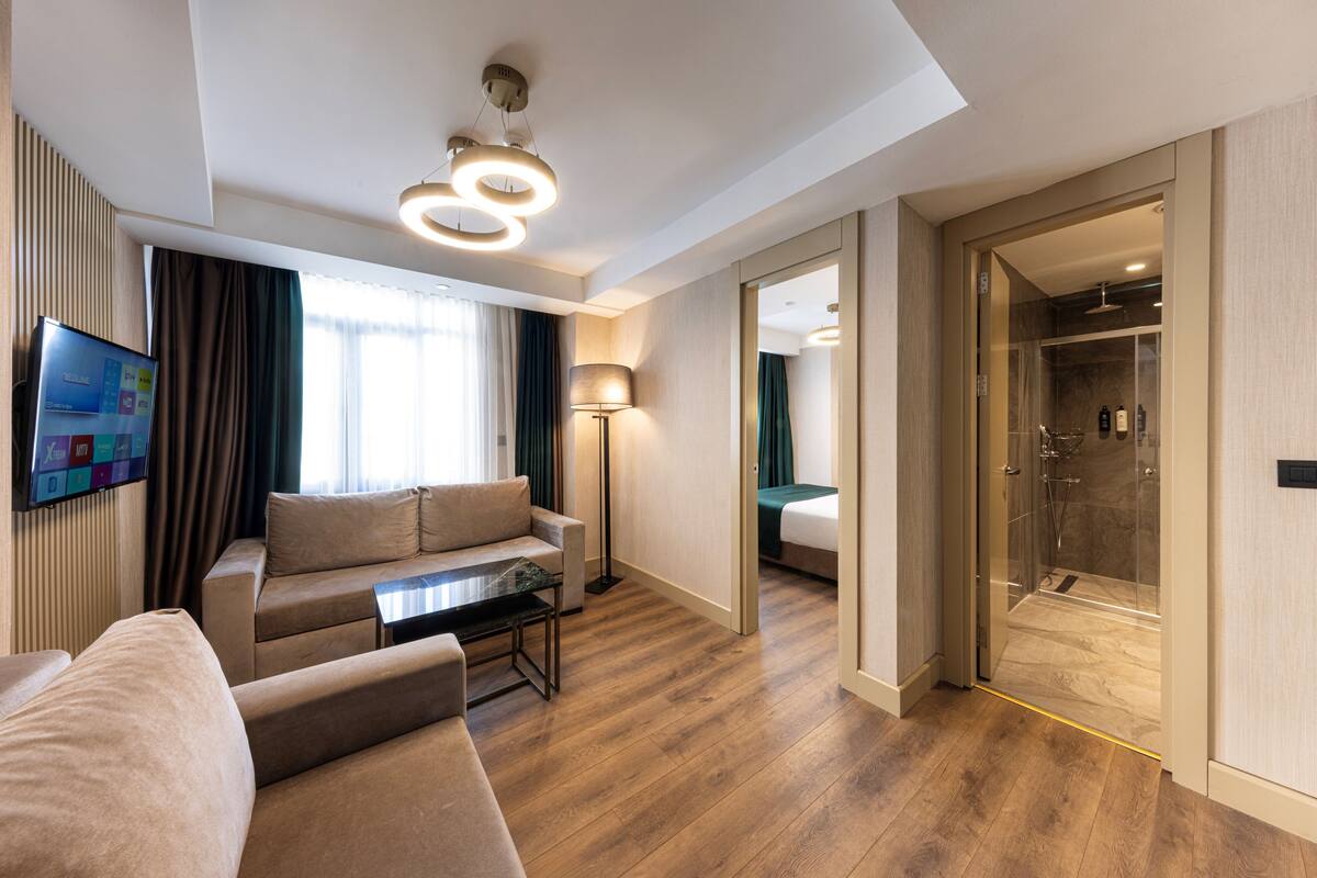 Una habitación de hotel moderna con sofá, una mesa de centro, televisor montado en la pared, lámpara de pie y cabina de ducha de vidrio, además de piso de madera.