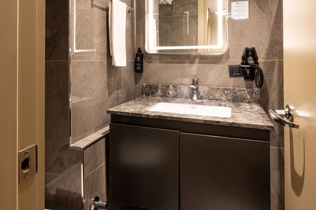 A modern bathroom with a marble sink, a large mirror with lights, a towel rack, and a hairdryer.