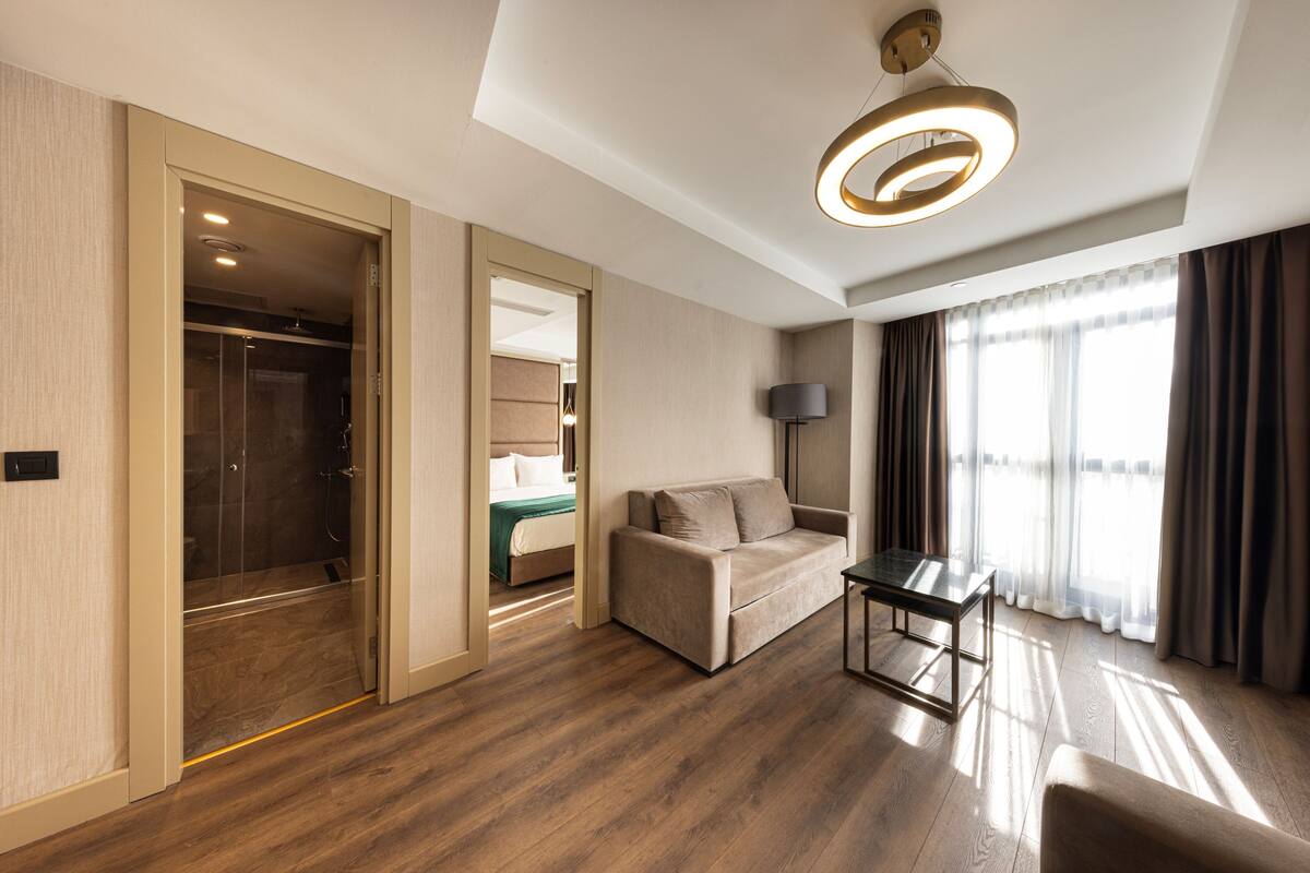 Habitación de hotel moderna con suelo de madera, un sofá, una mesa, una lámpara y vistas a un balcón.