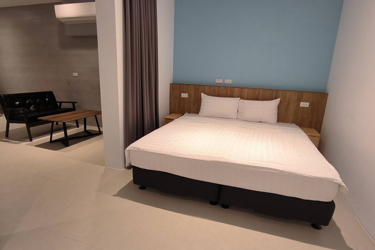 Una habitación de hotel moderna con una cama grande, un cabecero de madera, un banco y una cortina gris.