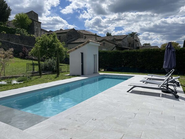Una piscina rectangular con una estructura pequeña adyacente, rodeada por un patio empedrado.