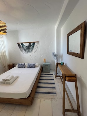 A bedroom with a wooden bed frame, a bedside table with a lamp, a small nightstand, a wall-mounted mirror, and a decorative wall hanging. - Luxurious Villa Oasis in Paros (Paros)
