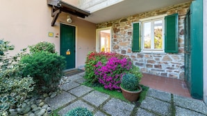 A stone building with a green door and shutters, a small garden with pink flowers, and a potted plant. - Solelago by Wonderful Italy (San Siro)