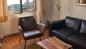 A living room with a view of a lake, a black leather sofa, a brown leather armchair, a wooden coffee table, and a floor lamp.