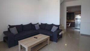 Un salon moderne avec un canapé d’angle aux couleurs sombres, une table basse en bois et un coin cuisine à l’arrière-plan.