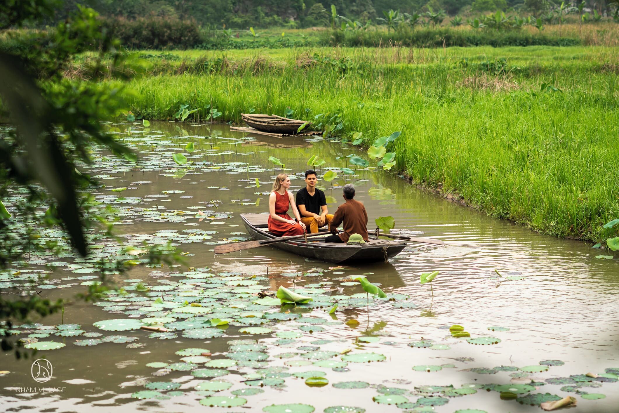 A boat with three people on a calm river surrounded by lush greenery and lily pads.