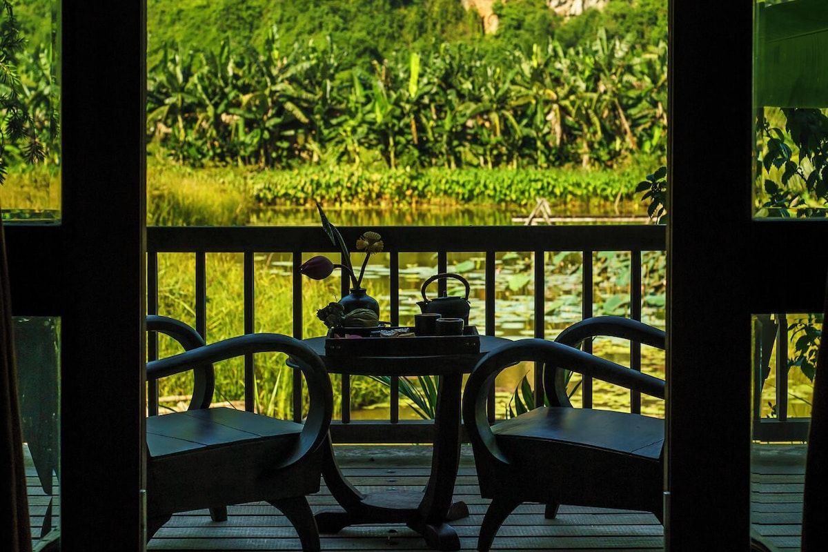 Una terraza de madera con dos sillas y una mesa, con vistas a un paisaje verde exuberante que incluye una masa de agua y una montaña.