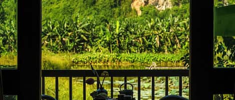 A wooden deck with two chairs and a table, overlooking a lush green landscape with a water body and a mountain.