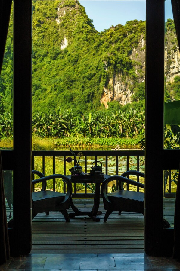 Una terraza de madera con dos sillas y una mesa, con vistas a un paisaje verde exuberante que incluye una masa de agua y una montaña.