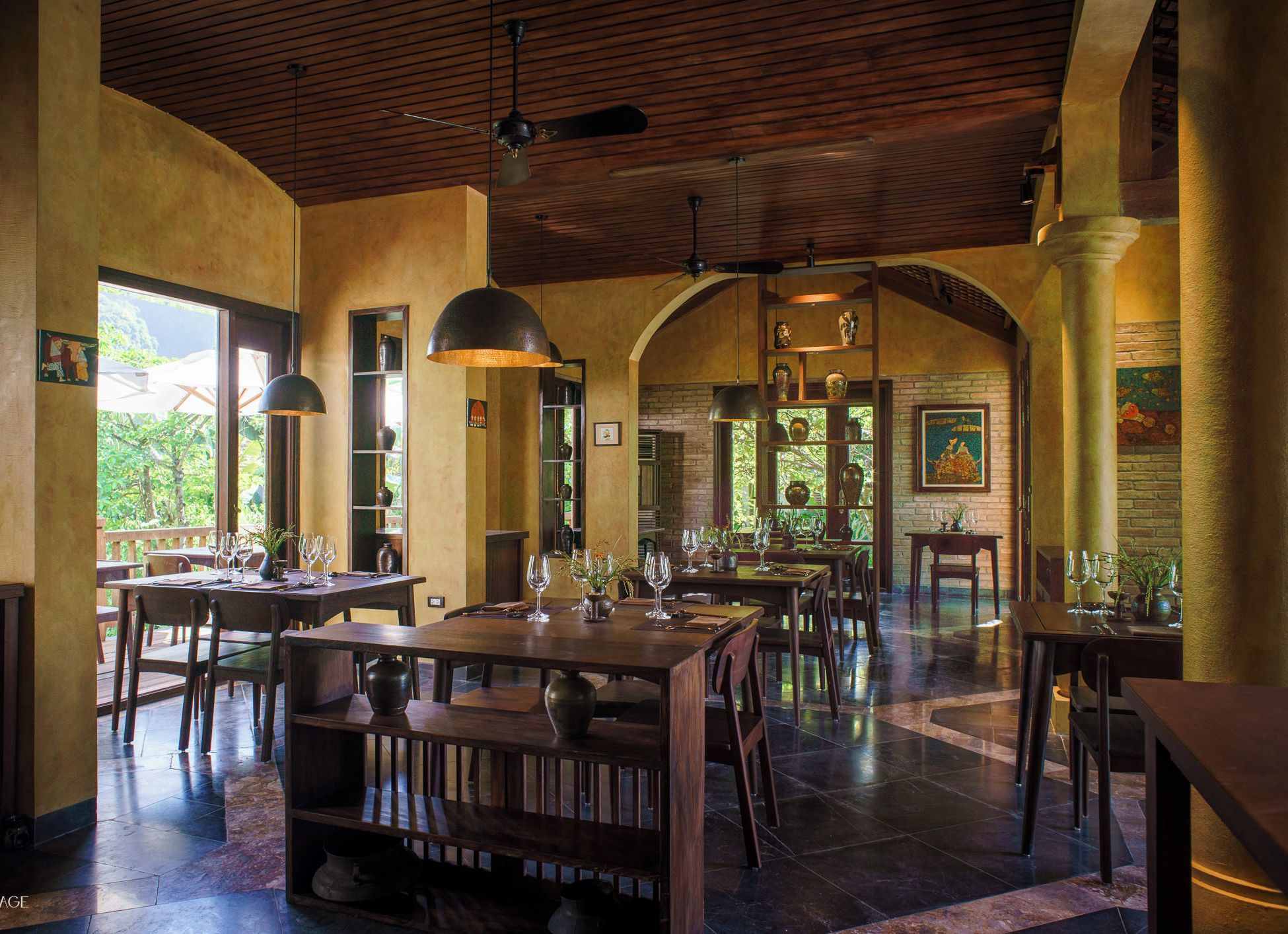 A spacious dining area with wooden tables and chairs, large pendant lights, and a view of greenery outside.