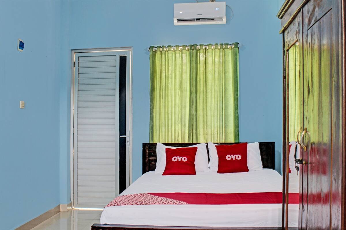 Ein Zimmer mit einem Bett, einer geschlossenen Tür, einem Fenster mit grünen Vorhängen und einer Klimaanlage an der Wand.