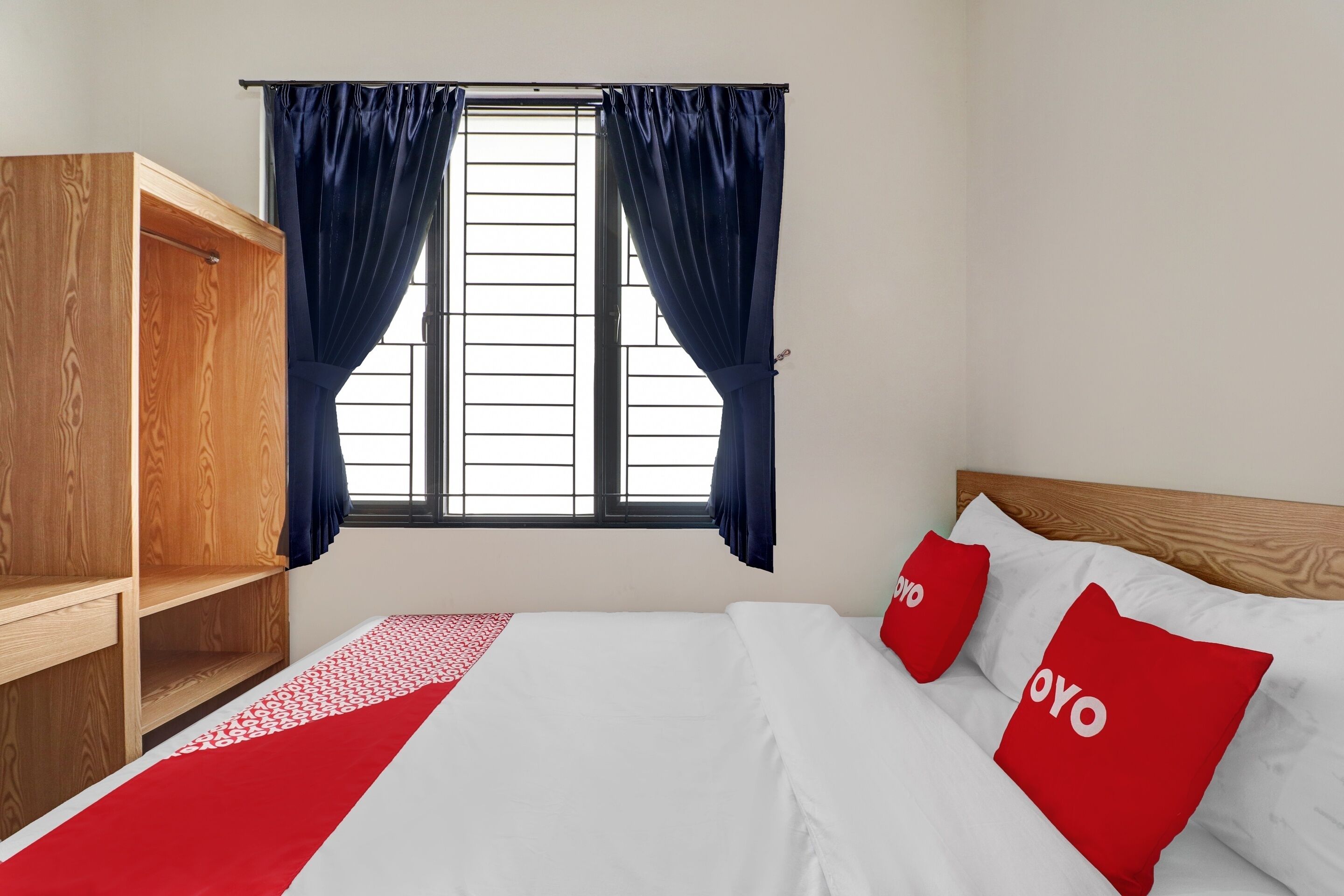 A bedroom with a wooden wardrobe, a bed with white and red bedding, and a window with blue curtains.
