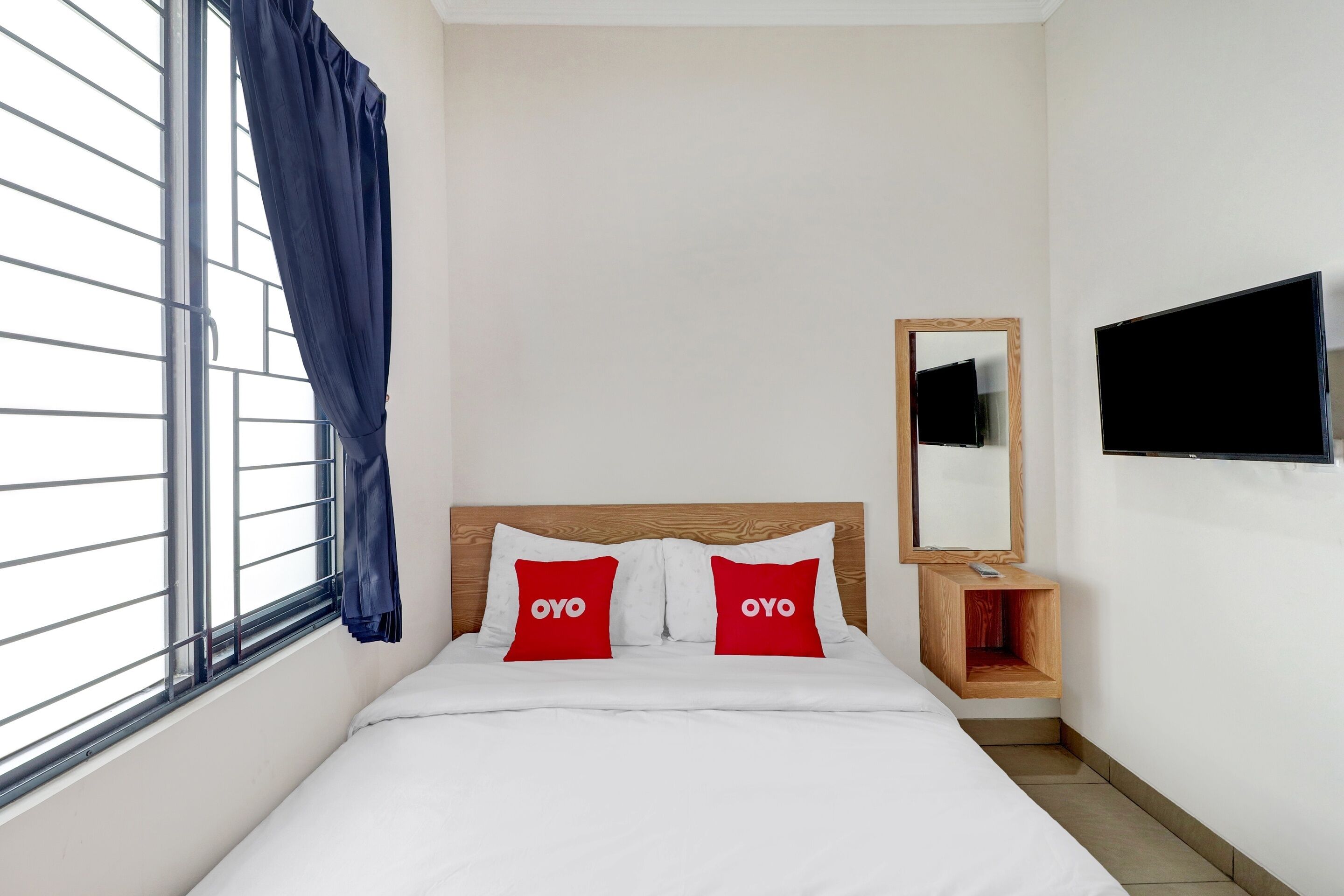 Camera d'albergo con un letto, due cuscini rossi con il marchio "OYO", una testiera in legno, una TV a parete e una finestra con tenda.