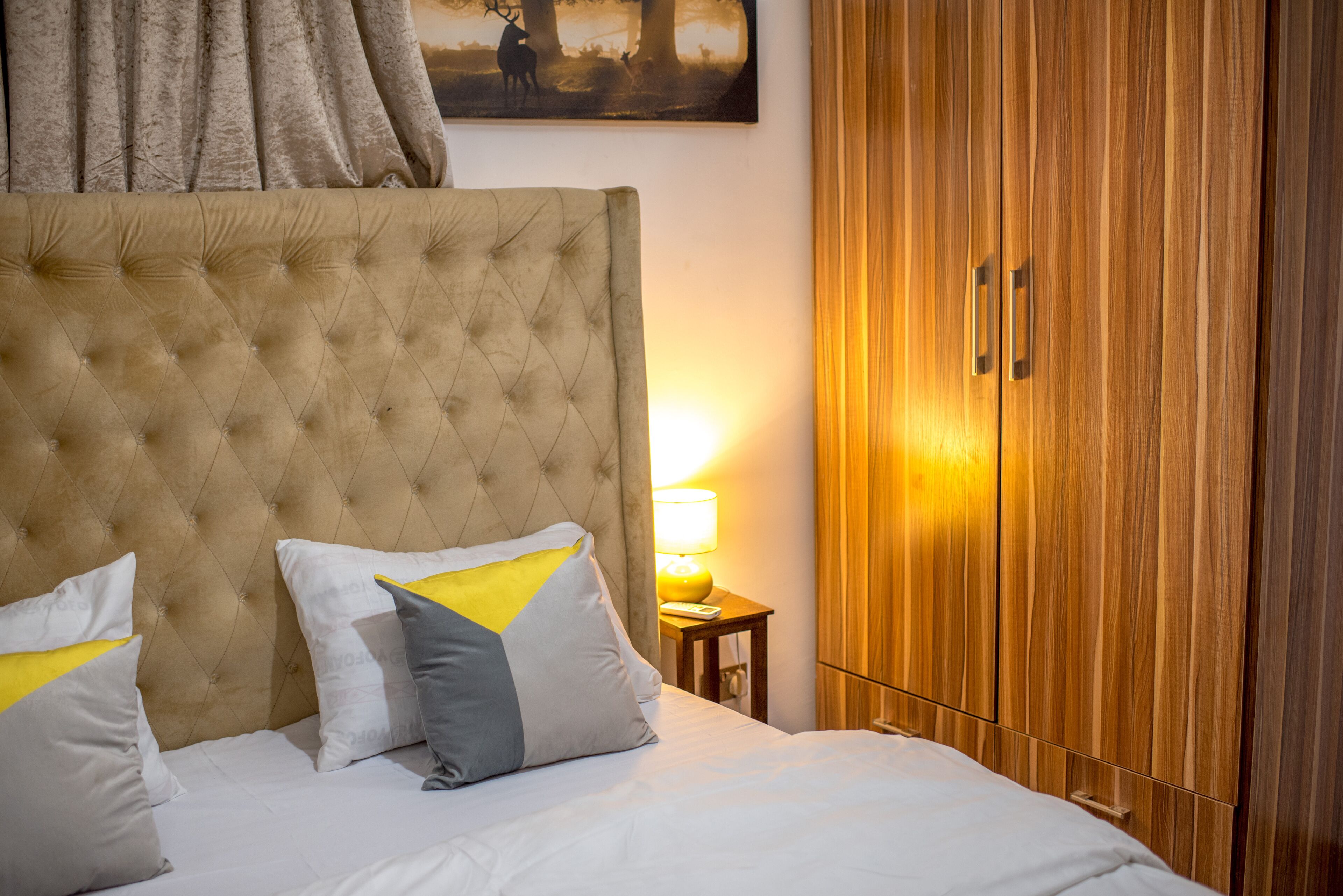 A hotel room with a tufted headboard, a bed with white linens and decorative pillows, a wooden wardrobe, and a painting of deer on the wall.