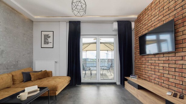 Una sala de estar moderna con un sofá, un televisor montado en una pared de ladrillo, una puerta corrediza de vidrio que da a un espacio al aire libre y una pequeña mesa con libros.