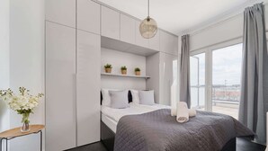 Un dormitorio moderno con cama, mesita de noche con un florero, ventana con cortinas y vista a la ciudad.