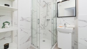 Een moderne badkamer met een douchecabine van glas, witte marmeren tegels, een witte wastafel en een spiegel met verlichting.