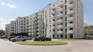 A multi-story residential building with balconies, a parking area, and a clear sky.