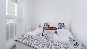 A bright, white room with a bed, a bedside table with a tray, and a window with sheer curtains.