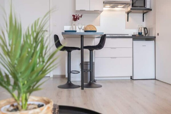 Una cocina moderna con armarios blancos, microondas y cafetera. Hay dos taburetes negros y una pequeña mesa con un florero.
