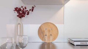 Una cucina moderna con mobili bianchi, un piano di lavoro nero e un tagliere di legno. Sul piano ci sono tre bicchieri da vino e un vaso con fiori rossi.
